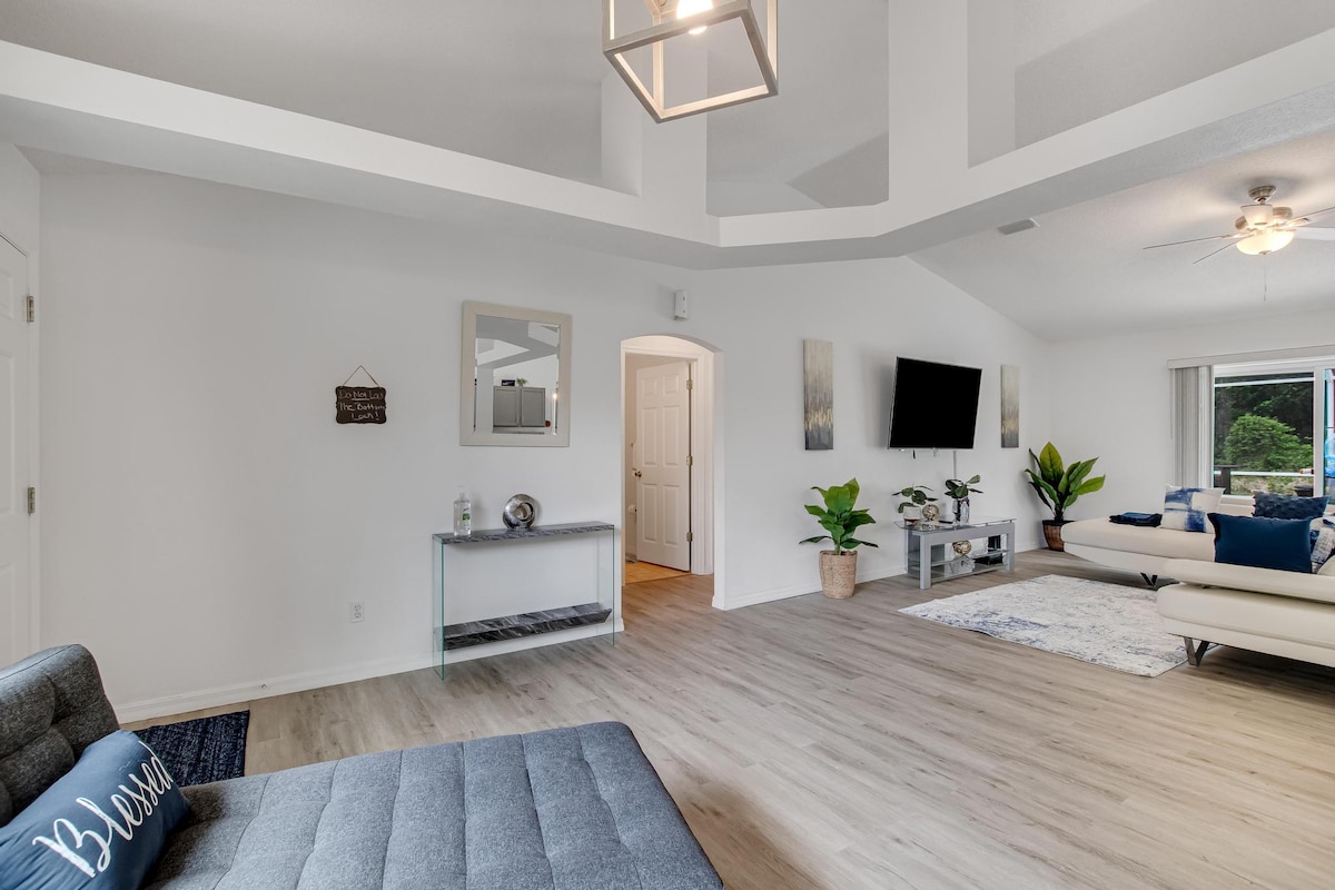 A spacious living area presents high ceilings and a contemporary ceiling fixture. A comfortable gray sofa is positioned adjacent to a large window, allowing natural light to illuminate the room. Decorative elements include a small console table and potted plants, enhancing the welcoming ambiance.