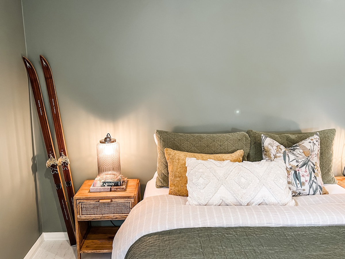 A spacious bed adorned with textured pillows and a soft blanket is positioned against a green wall. A bedside table holds a decorative lamp, while vintage wooden skis are leaning against the wall, adding an outdoor element to the interior.