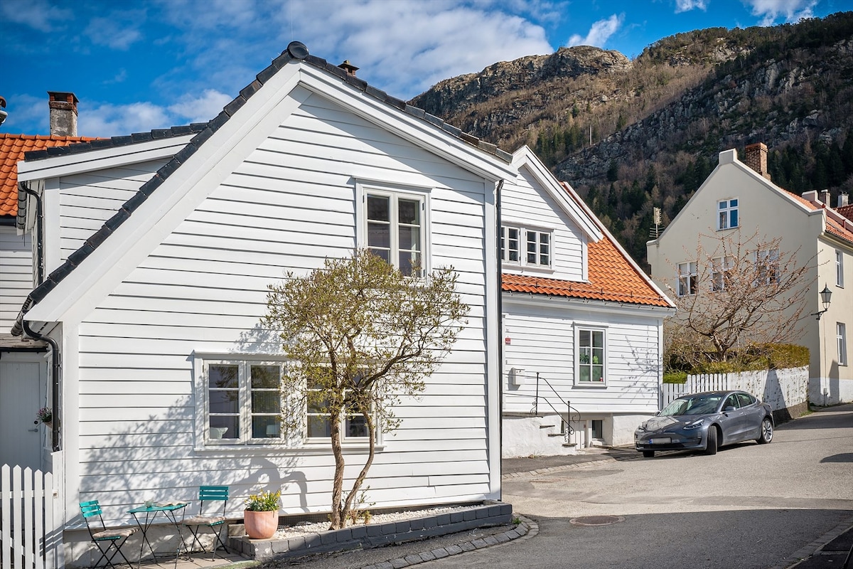 A charming white wooden house from the 1700s is shown, reflecting a blend of historic and modern elements after renovation. A small tree is visible in front, while two teal chairs are positioned on a patio area. The backdrop features a hillside and additional nearby buildings.