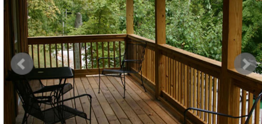 A wraparound deck is lined with wooden railings and features several black metal chairs, offering a casual seating area. The wooden floor is complemented by surrounding greenery visible through the openings in the railings.