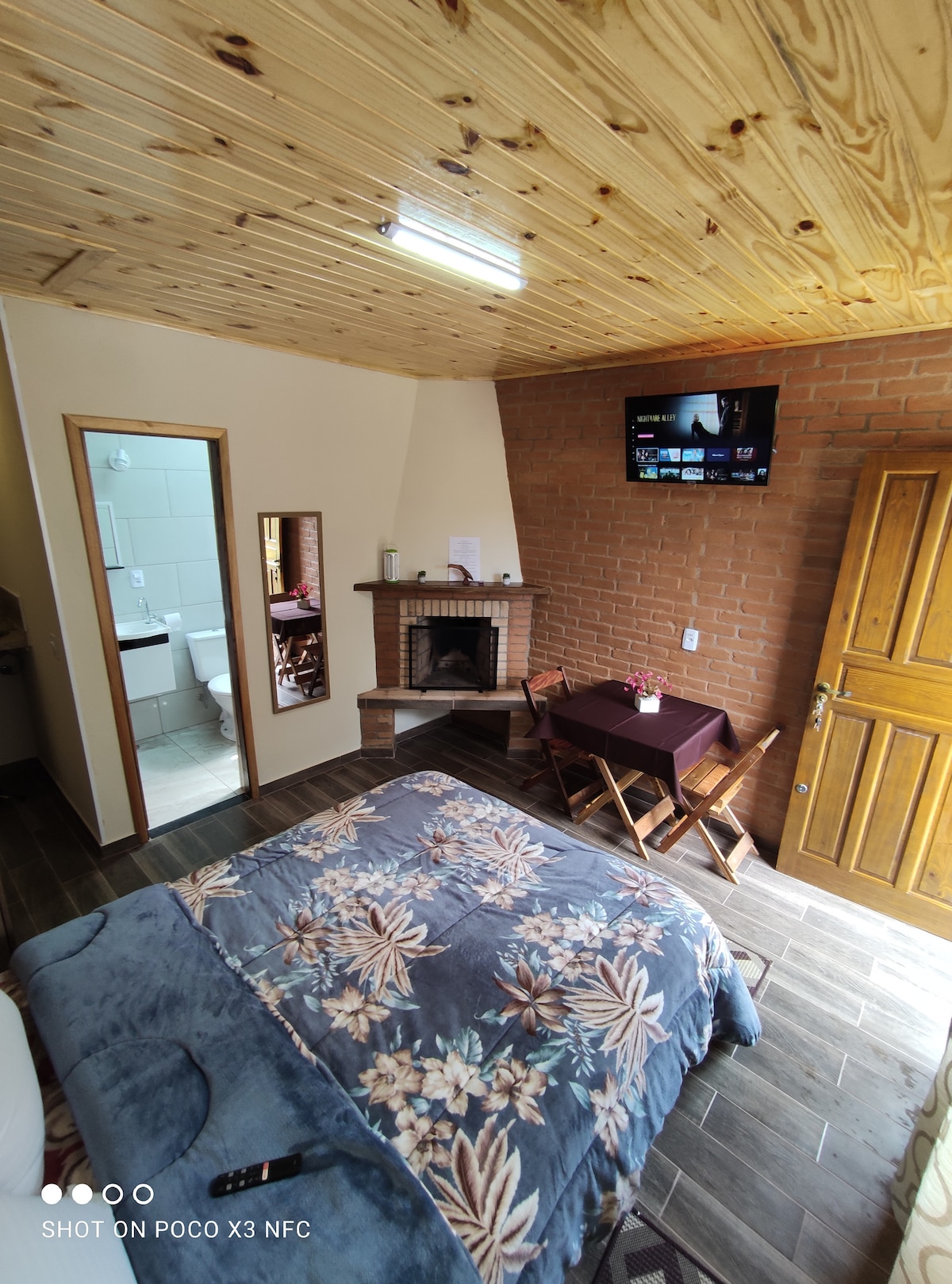 A cozy room features a spacious bed covered with a floral blanket. A fireplace is located against a brick wall, with a television mounted above it. A small table with two chairs is positioned near a mirror, enhancing the room's functional layout.