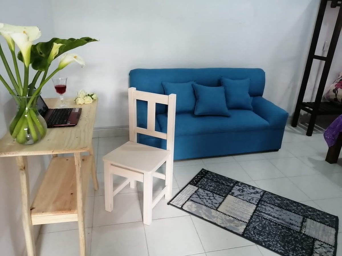 A small living area is shown with a blue sofa adorned with cushions. A wooden table is positioned beside it, featuring a laptop and a glass. A single white chair is placed next to the table on a patterned rug, enhancing the space's functionality.