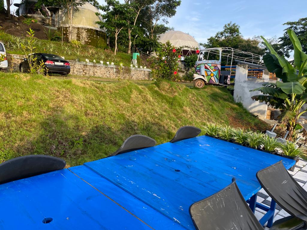 A vibrant blue table is positioned outdoors, accompanied by several dark chairs. The surrounding grassy slope is adorned with lush greenery and flowering plants, with parked vehicles visible in the background. A decorative vehicle adds character to the serene outdoor setting.
