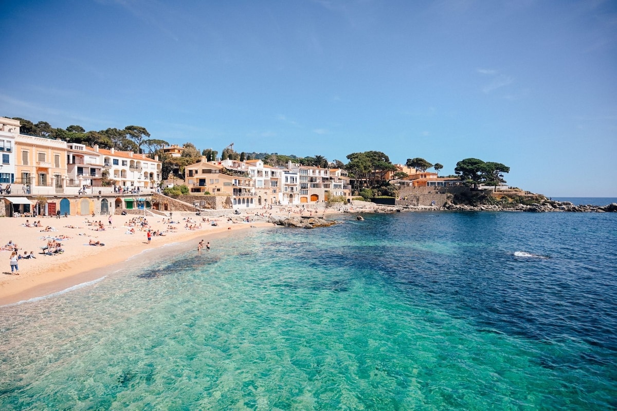A serene beach scene shows soft sands merging with clear turquoise waters. Colorful buildings line the shoreline, and sunbathers relax on the beach. Gentle waves lap at the shore, while greenery can be seen in the distance, contributing to the tranquil coastal ambiance.