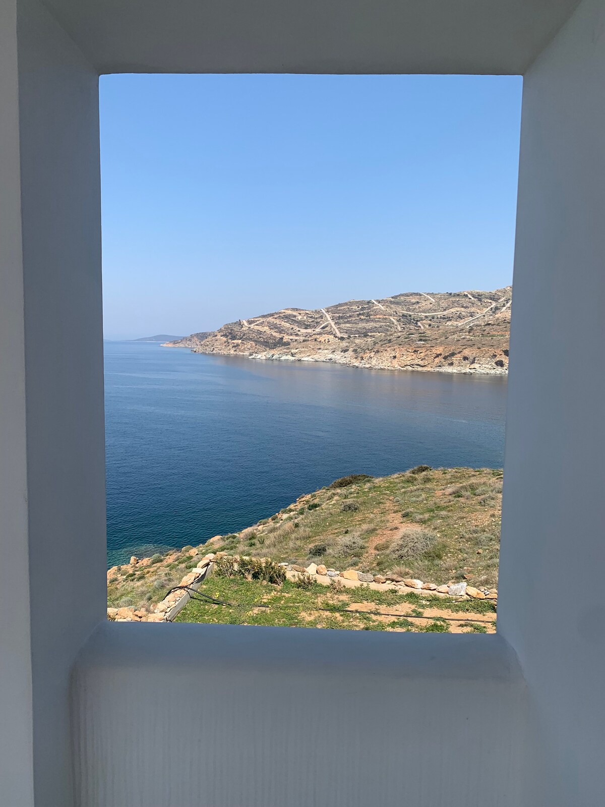 A scenic view is framed by a white structure, showcasing a tranquil coastline with gently rolling hills. The clear blue sea meets the horizon under a bright sky, providing a peaceful backdrop that highlights the natural beauty of the surrounding landscape.