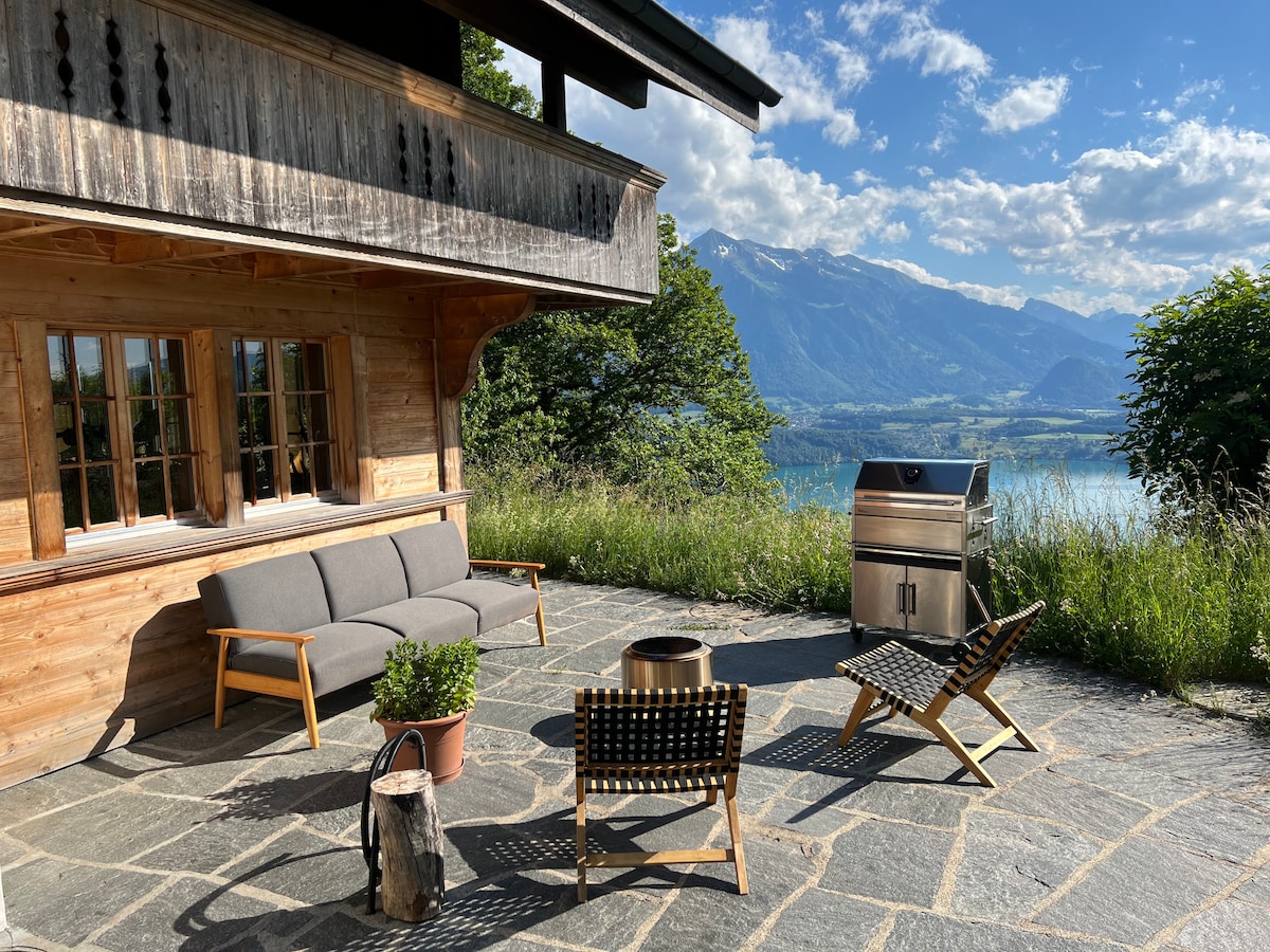 An outdoor seating area features a gray sofa and two wooden chairs arranged around a fire pit, all situated on a stone patio. A stainless steel grill is present nearby, with lush greenery and a scenic view of the lake and mountains in the background.