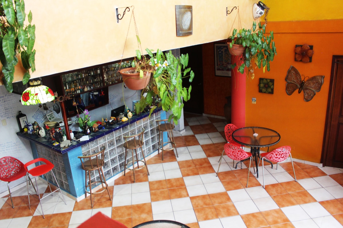 A communal area is presented with colorful walls and terracotta tile flooring. The space features hanging plants and a blue bar counter adorned with various beverages. Red chairs and glass-top tables are arranged for seating, providing a welcoming environment for guests.