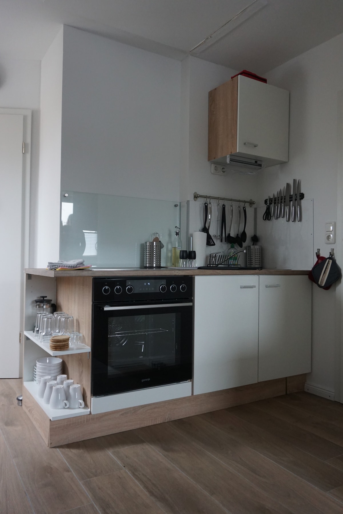 The image shows a compact kitchen area featuring a modern oven with a glass top and a sleek white cabinetry. Cooking utensils are organized neatly on a wall-mounted rack, while a glass panel provides an open feel. Shelves contain a selection of glassware and cooking essentials.