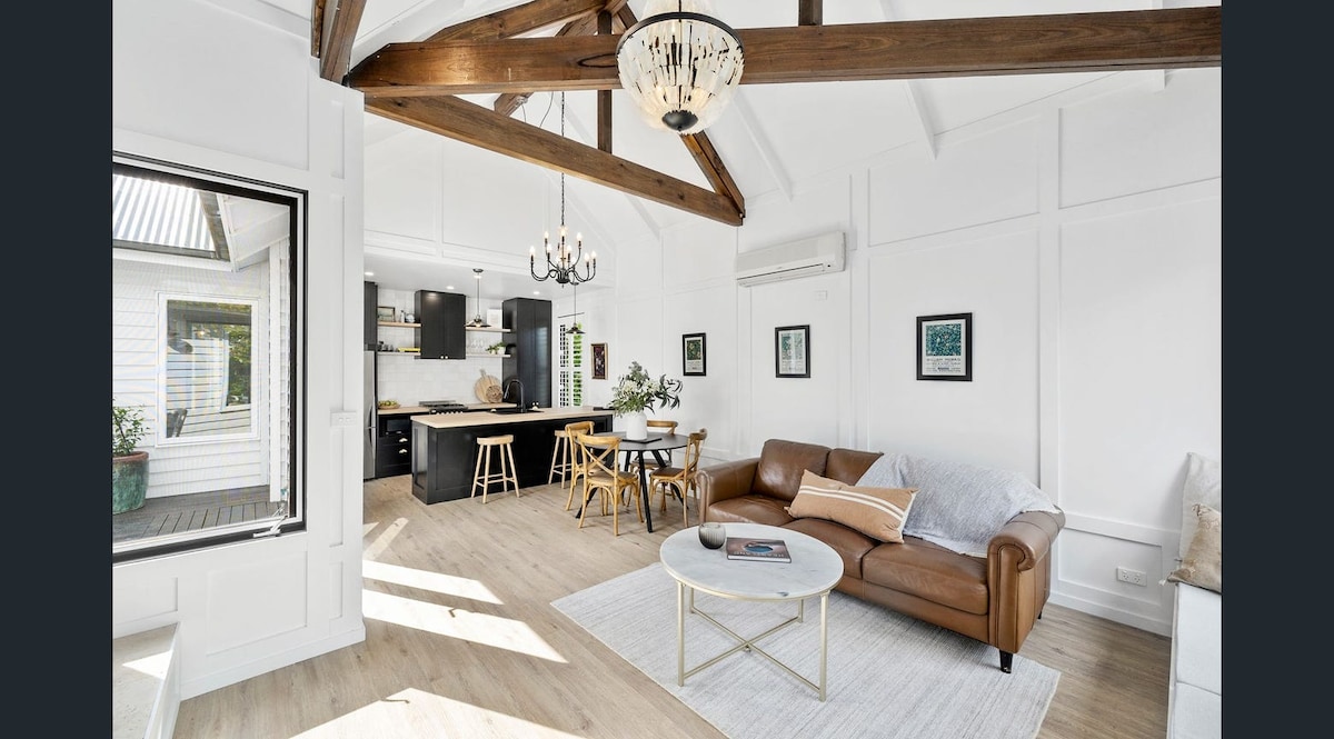 An open living area features vaulted ceilings supported by exposed timber beams, creating an airy atmosphere. A leather sofa is positioned across from a round coffee table. A dining area with wooden chairs and a stylish kitchen is visible in the background, complemented by natural light from nearby windows.