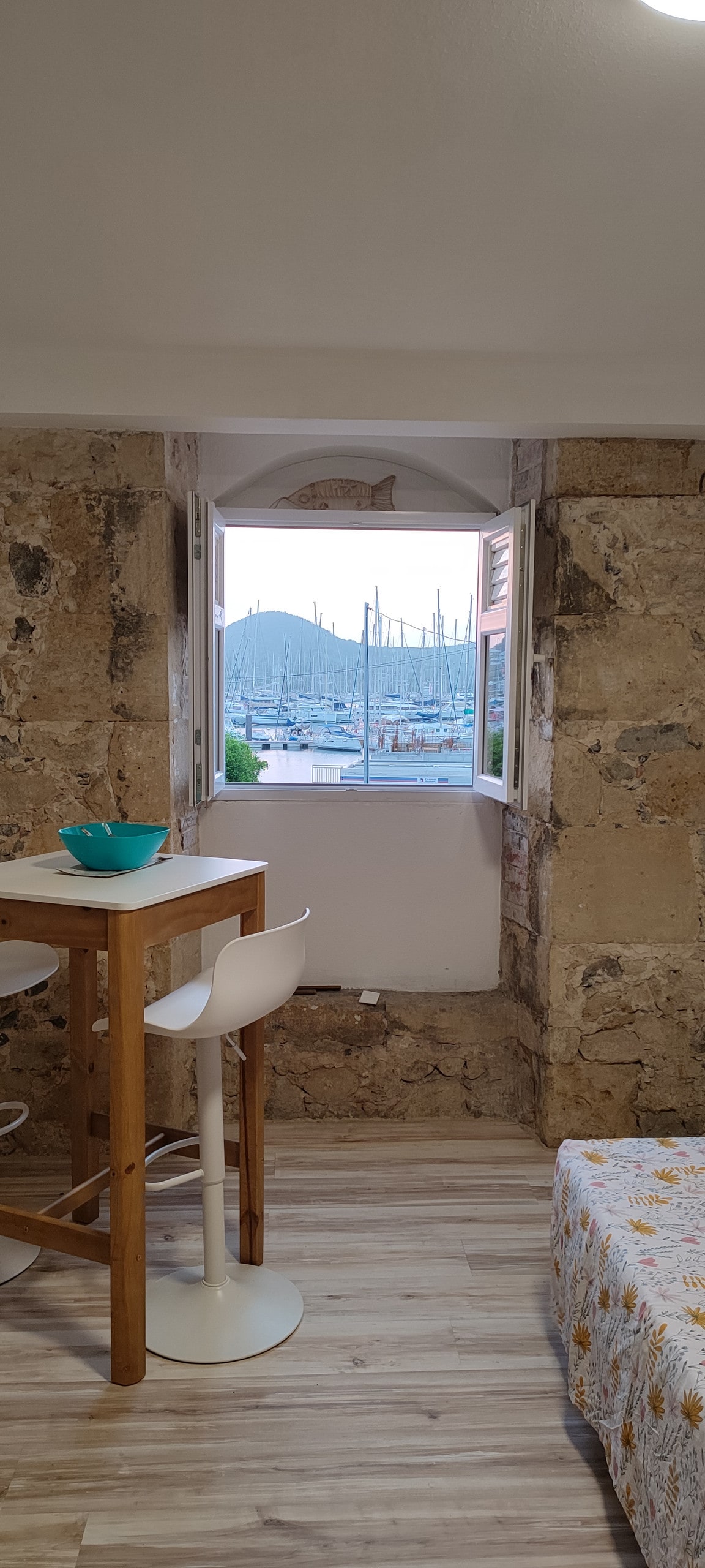 A window reveals a view of the marina, showcasing boats gently bobbing on the water. Natural light fills the space, illuminating the stone walls. A small dining table with stools is positioned nearby, contributing to a relaxed ambiance.