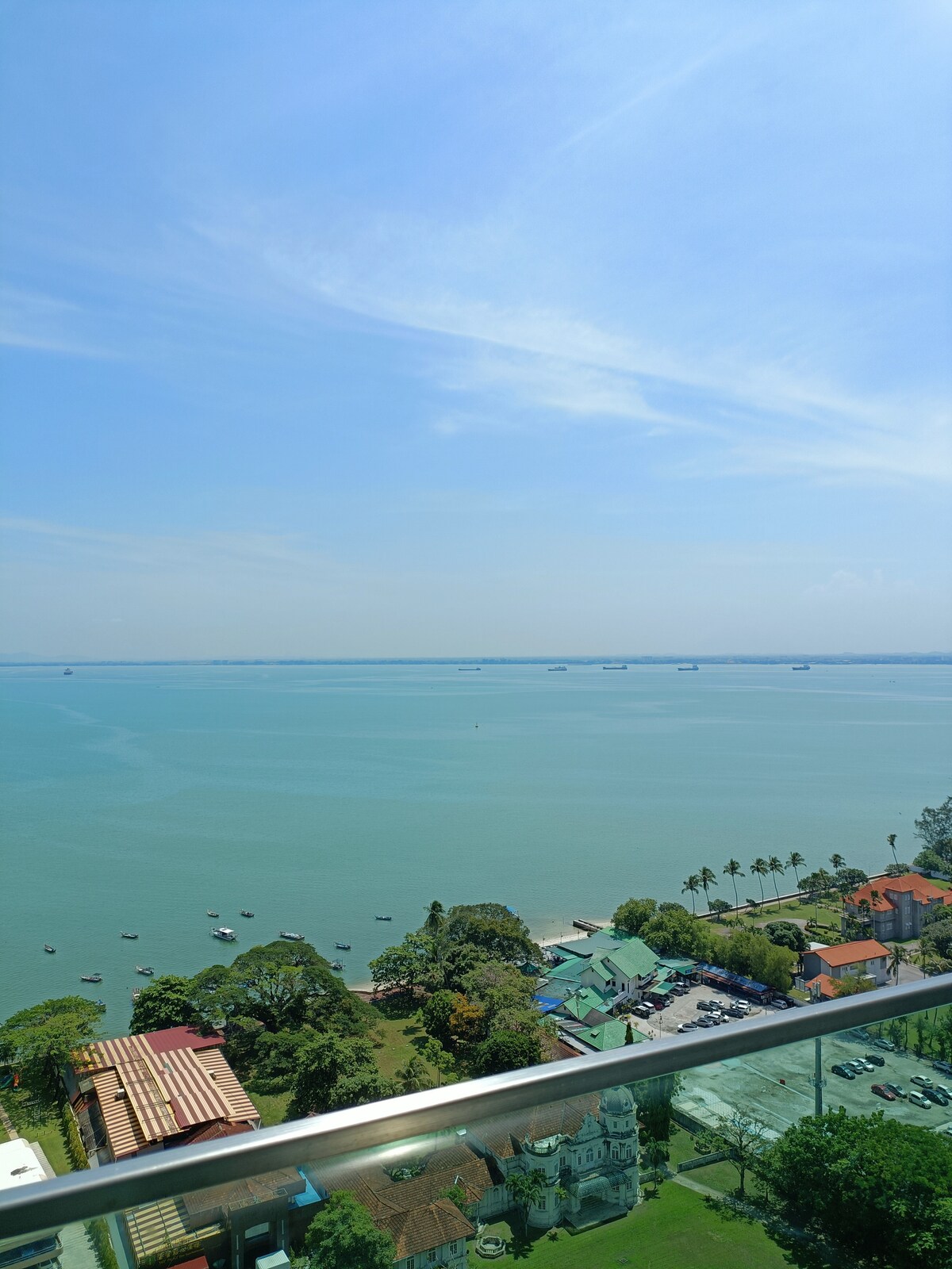 A panoramic view of the waterfront is displayed, with calm blue waters extending towards the horizon. A mix of lush greenery, palm trees, and residential buildings can be seen along the shore, while distant vessels dot the water under a clear blue sky.