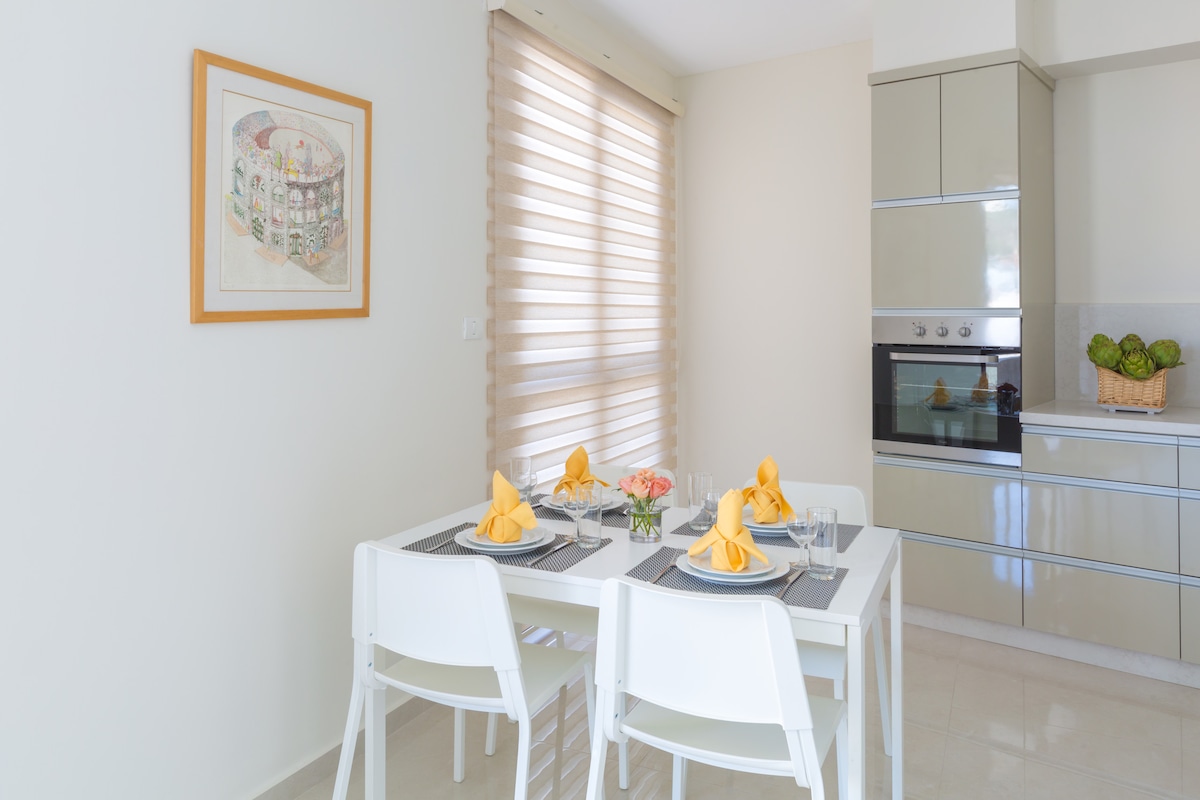 A dining area is presented with a white table set for four, featuring neatly folded yellow napkins and stylish glassware. Soft shades cover the windows, allowing natural light to brighten the space. A modern kitchen with sleek cabinetry and an oven is visible in the background.