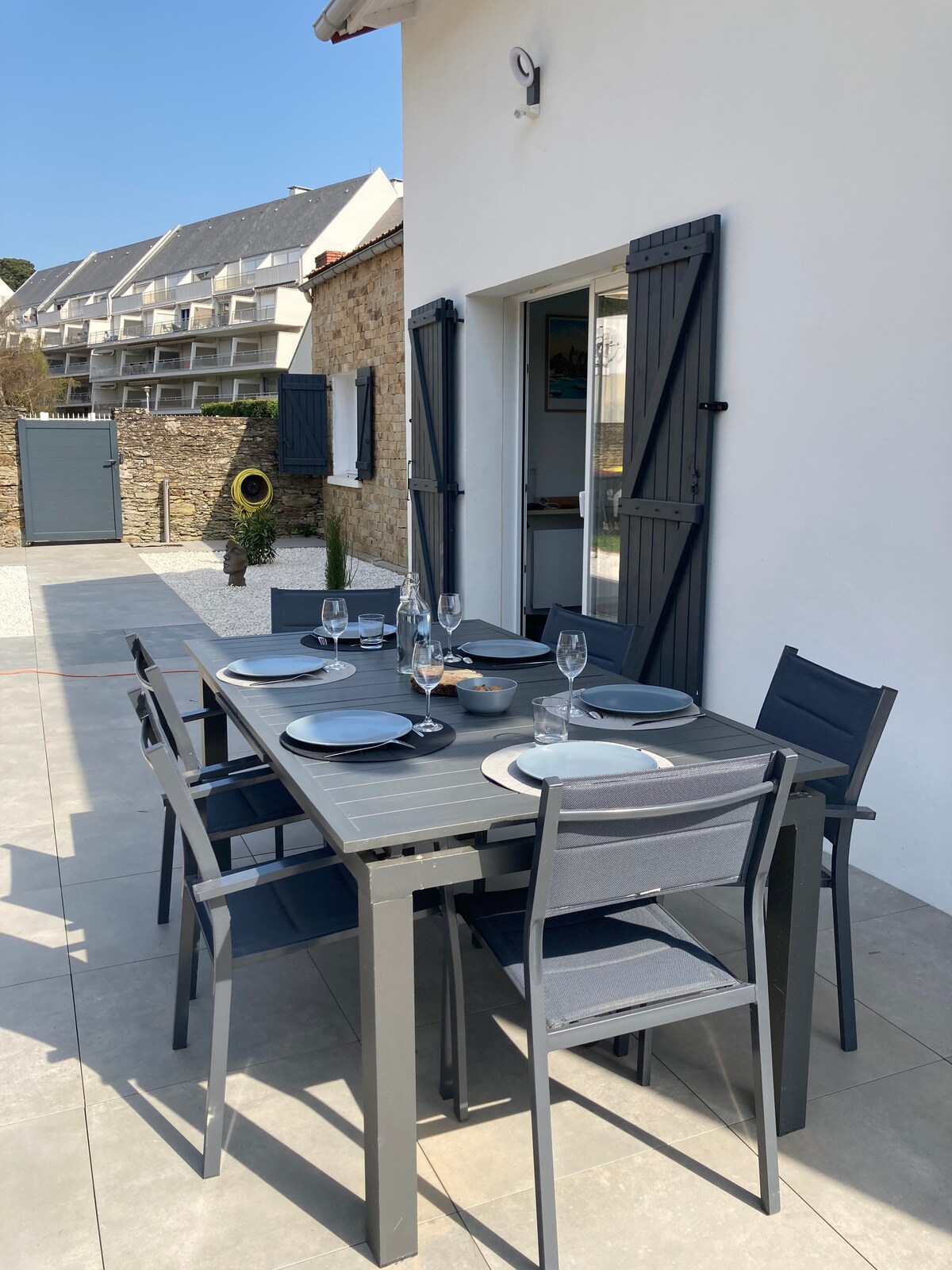 A dining area is set outdoors with a large gray table and six matching chairs. Plates, glasses, and silverware are neatly arranged on the table. The entrance is framed by grey shutters, adding a modern touch to the white wall.