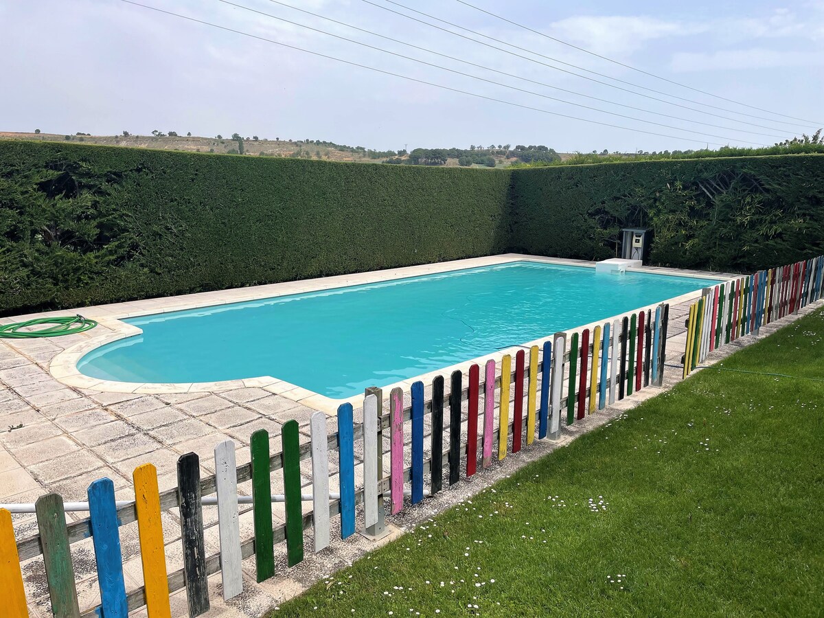A large swimming pool features bright turquoise water, surrounded by a manicured lawn and a low fence painted in various colors. Tall green hedges provide privacy along the perimeter, with a clear sky visible above.