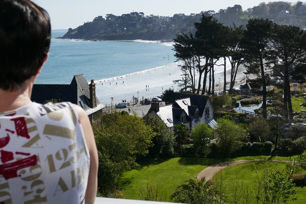 A view from the terrace showcases a sandy beach with gentle waves and people enjoying the seaside. In the foreground, lush greenery surrounds quaint buildings, while distant hills frame the picturesque coastline under a clear sky.