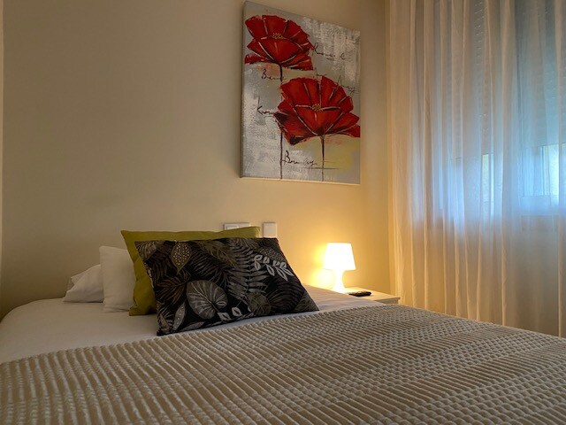 A bright sleeping area features a spacious bed adorned with a textured blanket and decorative pillows. A bedside lamp provides soft lighting next to the bed. A floral artwork in red hangs above, with light filtered through sheer curtains from the window.