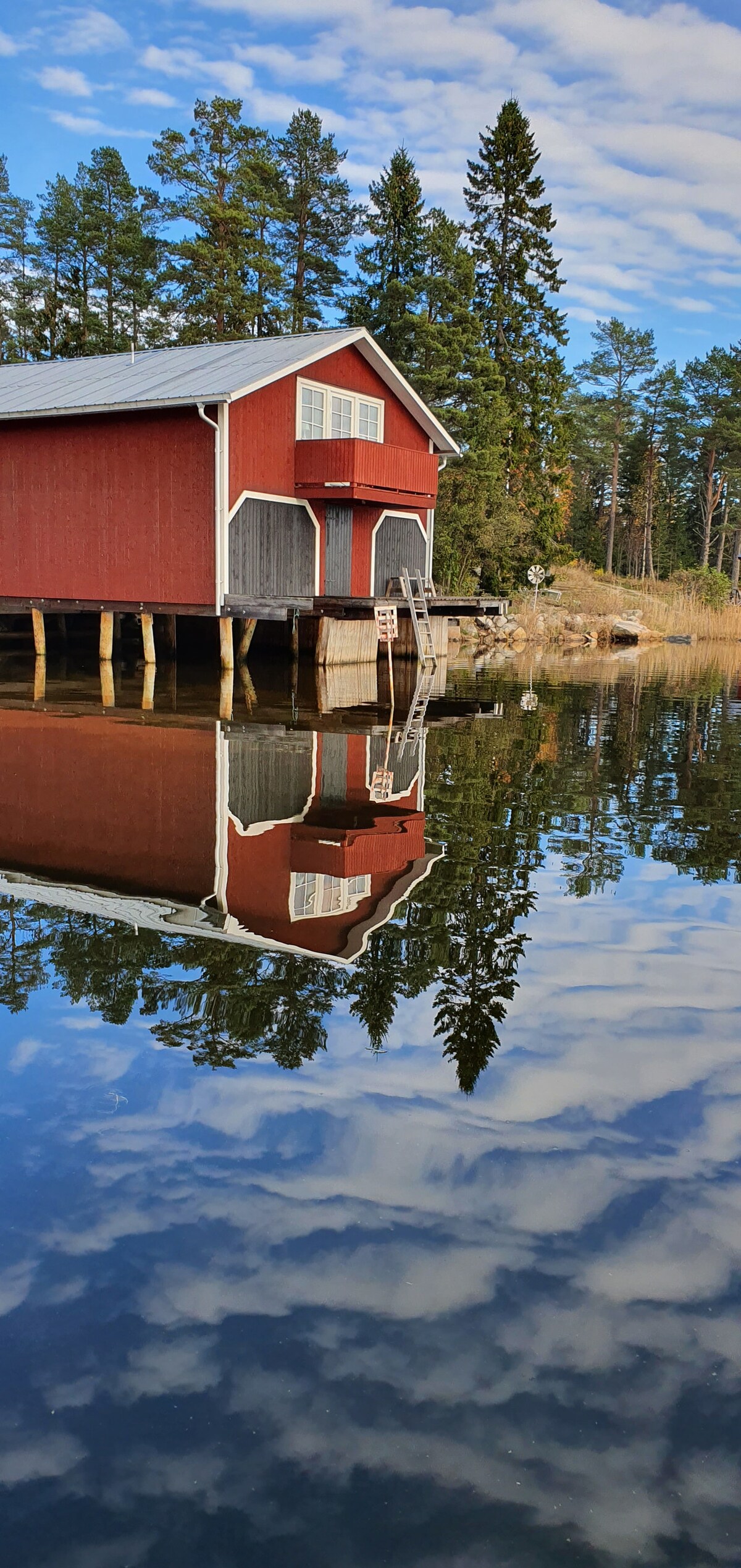 A red wooden building is situated by the water's edge, reflecting in the calm water below. Surrounding trees and a partly cloudy sky are also mirrored in the water, creating a serene atmosphere. The structure features white accents and sits on stilts.