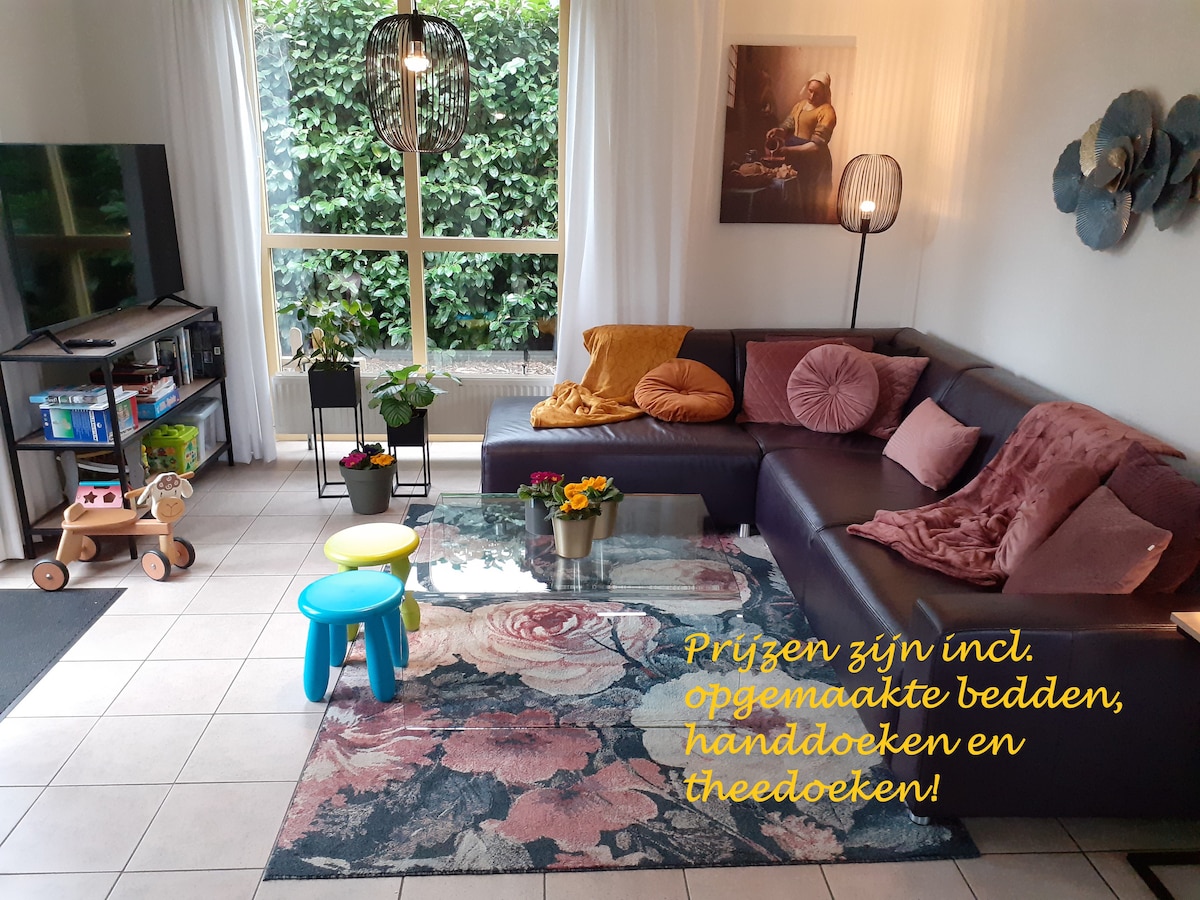 A comfortable living area features a dark sectional sofa with pillows, a colorful area rug adorned with floral patterns, and a glass coffee table centered with a small flower arrangement. Large windows allow natural light, while a shelving unit and a play area are visible in the background.