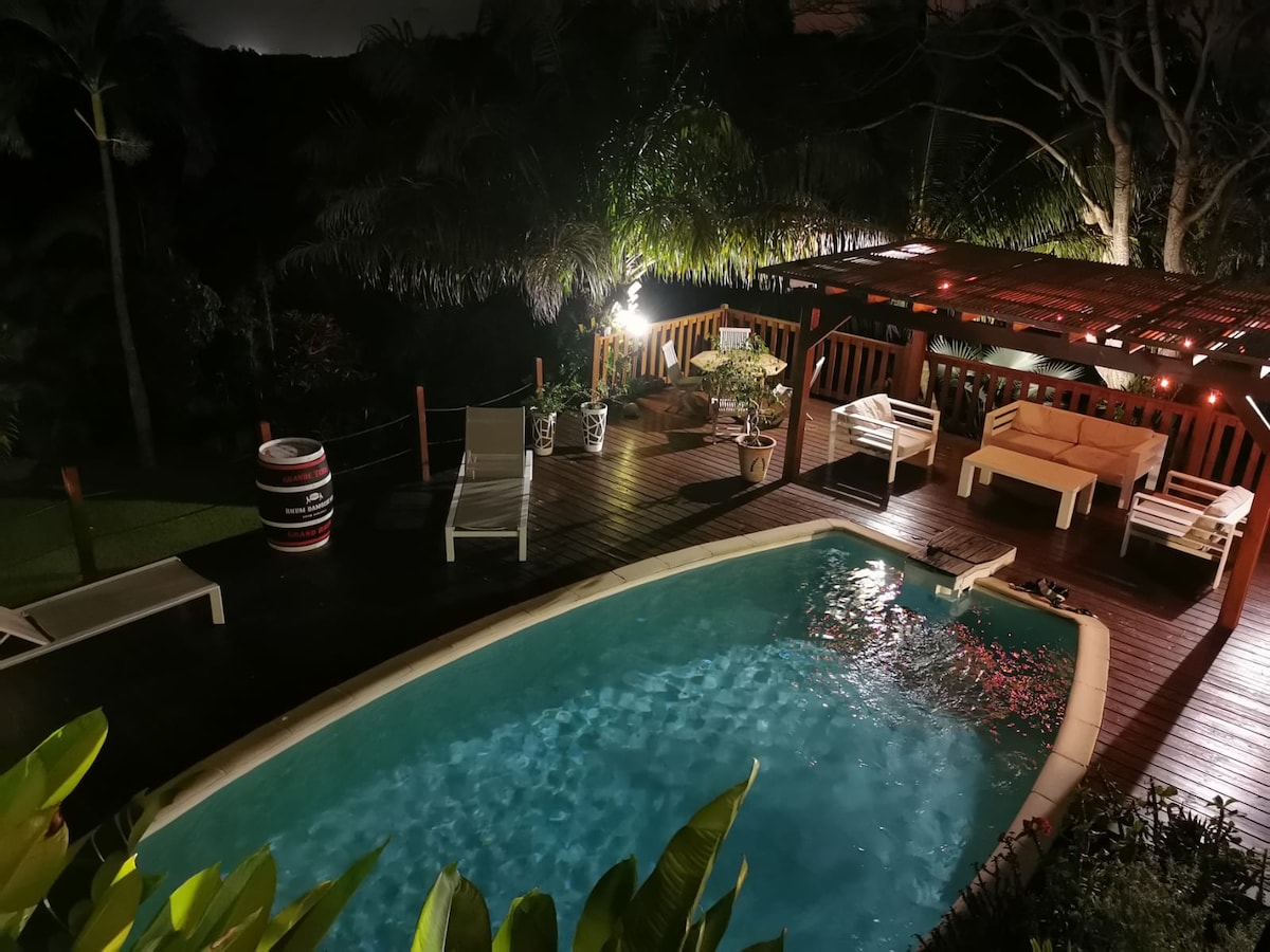 A serene outdoor space features a swimming pool with clear water, surrounded by lounge chairs and a wooden deck. Potted plants adorn the area, while a cozy seating arrangement sits beneath a pergola, illuminated softly by surrounding lights.
