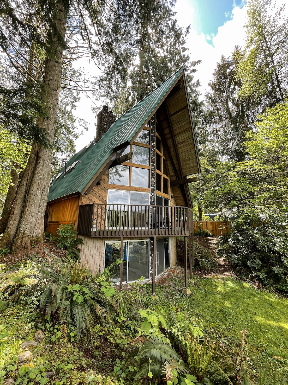 The exterior of the A-frame cabin is framed by lush greenery and towering trees. A three-story wall of windows allows natural light to flood in, while a wooden balcony extends out, inviting guests to enjoy the natural surroundings.