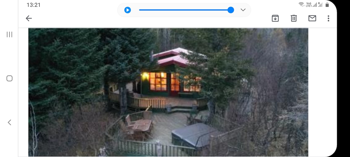 Aerial view of a cozy cabin surrounded by trees, featuring large windows that allow natural light to fill the interior. A wooden deck extends around the cabin, with a hot tub positioned nearby for relaxation.