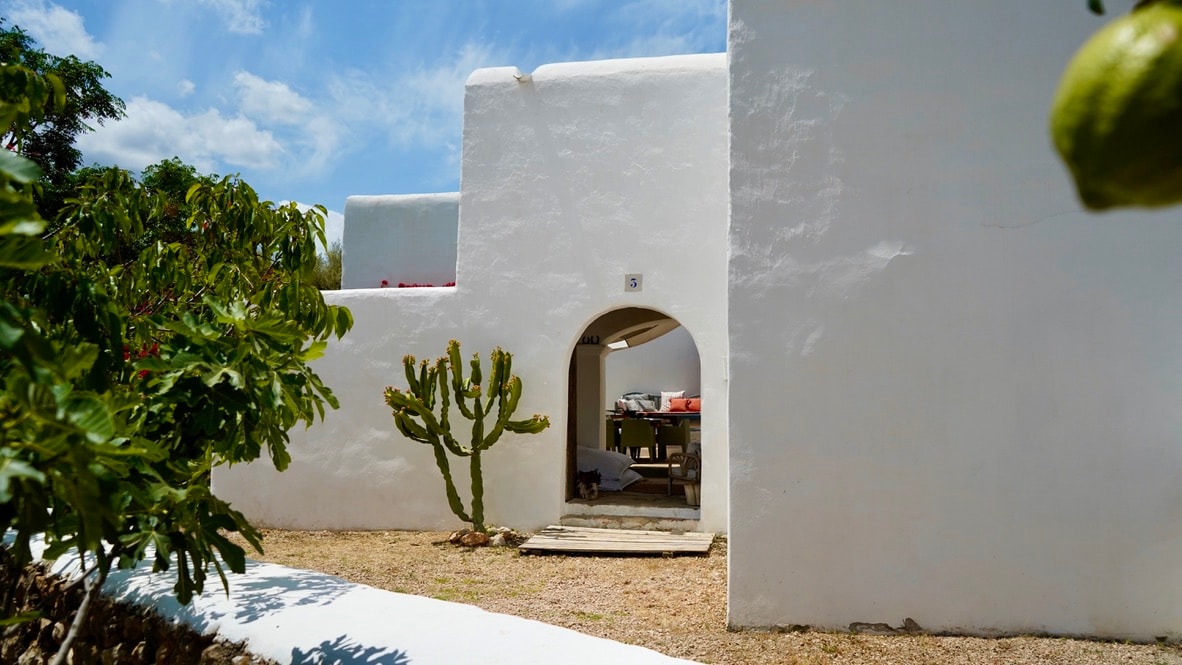 The white-washed exterior of the finca is framed by green foliage, including a cactus in the foreground. An arched entrance leads to a shaded outdoor dining area, with rustic wooden furniture visible inside. The bright sky adds a serene atmosphere.