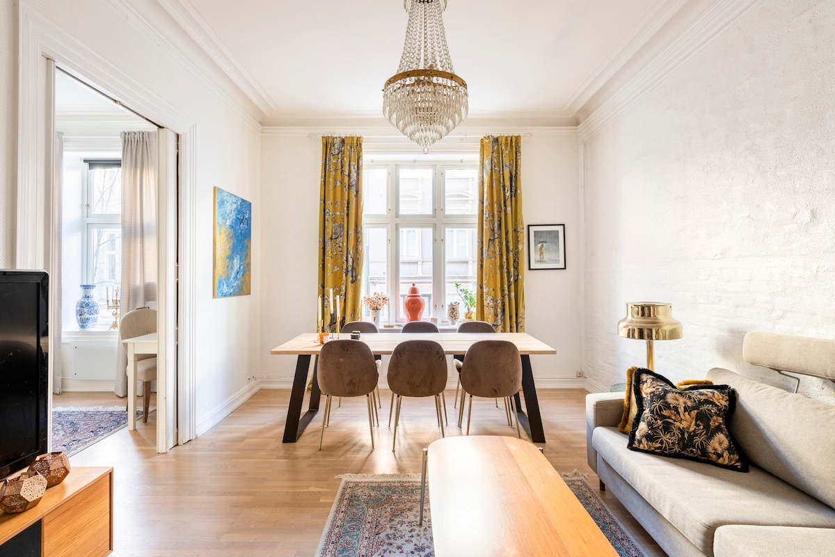 A dining area features a long table surrounded by six chairs, illuminated by natural light from large windows with yellow curtains. A cozy seating arrangement with a sofa is visible, along with decorative elements like wall art and a plush rug.