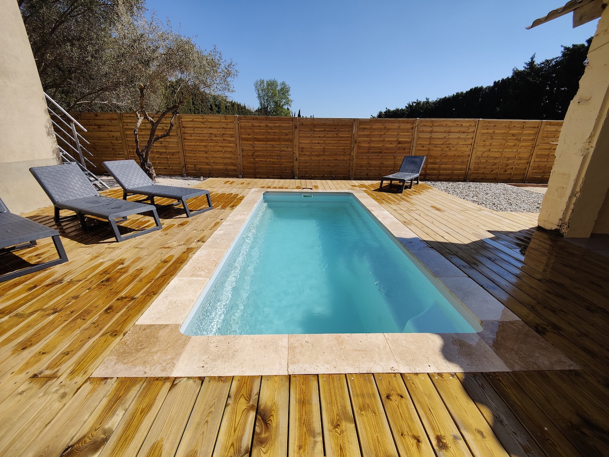 A private pool is set within a wooden deck, surrounded by sun loungers. The tranquil water reflects clear blue skies, and a wooden fence offers privacy. Nearby, olive trees provide a natural backdrop, enhancing the serene outdoor space.