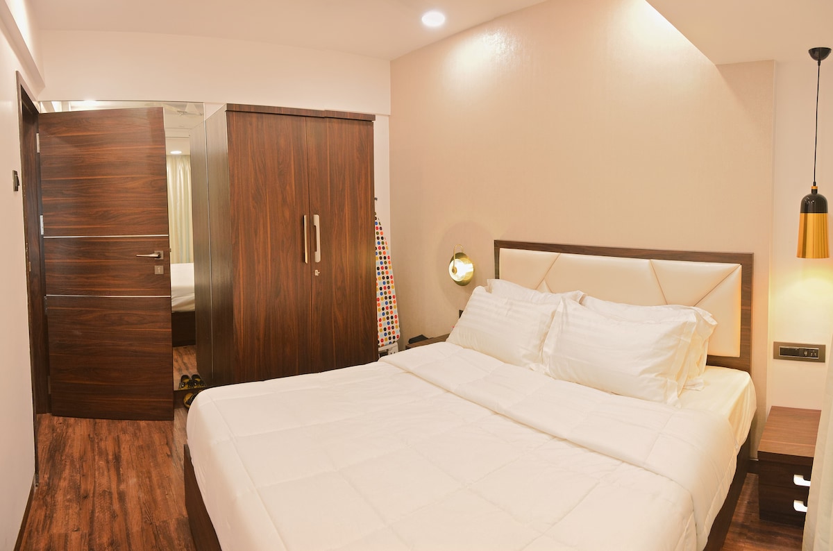 A well-furnished bedroom is presented with a spacious bed featuring crisp white linens. A wooden wardrobe stands nearby, complemented by a contemporary wall light. A mirrored door reflects additional room space, enhancing the functionality and design of the area.