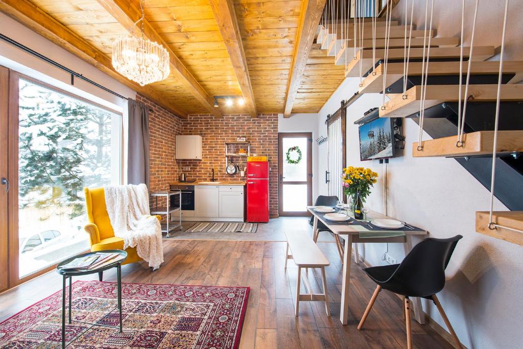 An open living space features a bright yellow armchair and a wooden dining table set with black seating. Exposed brick walls complement the wooden beams overhead. A decorative area rug adds warmth, while a vibrant red refrigerator enhances the kitchen area, illuminated by natural light.