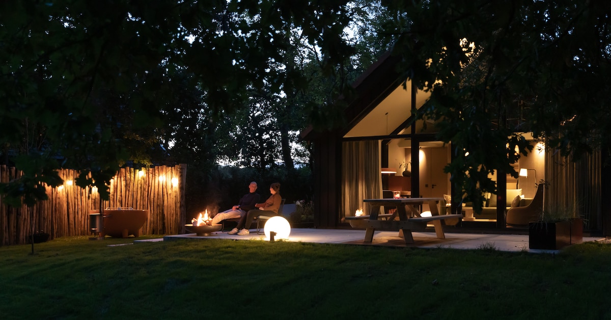 The cabin is illuminated softly in the evening, surrounded by trees. A wooden picnic table and a circular light fixture are displayed on the spacious terrace. Two chairs are positioned beside a small fire, creating a cozy outdoor seating area under the canopy of twilight.