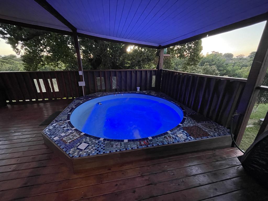 A circular hot tub is set on a wooden deck, surrounded by a mosaic tile border. Soft blue lighting reflects off the water, creating a calming ambiance. The area is shaded by a roof, with greenery visible in the background.