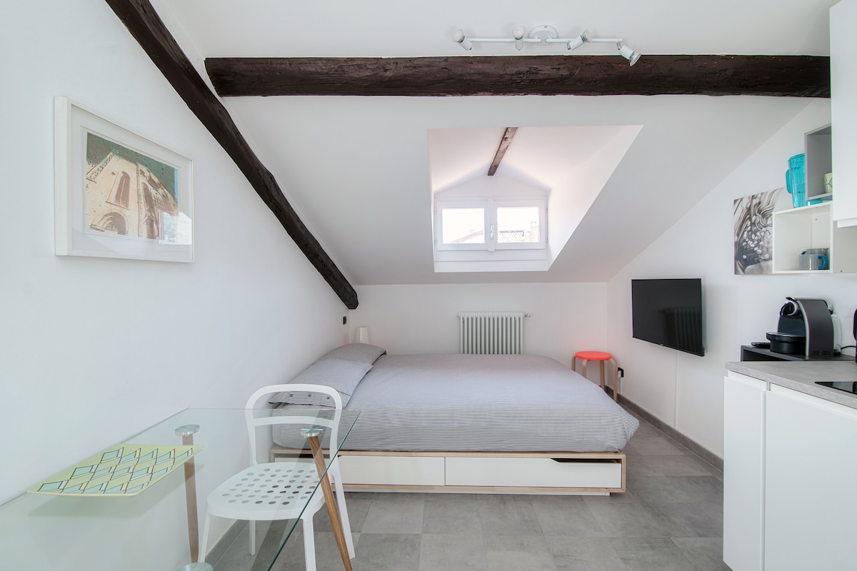 A cozy bedroom features a spacious double bed with light grey bedding. A skylight brightens the room, illuminating the white walls and exposed wooden beams. A small glass table with a translucent chair complements the minimalist decor, alongside a compact kitchenette and wall-mounted television.