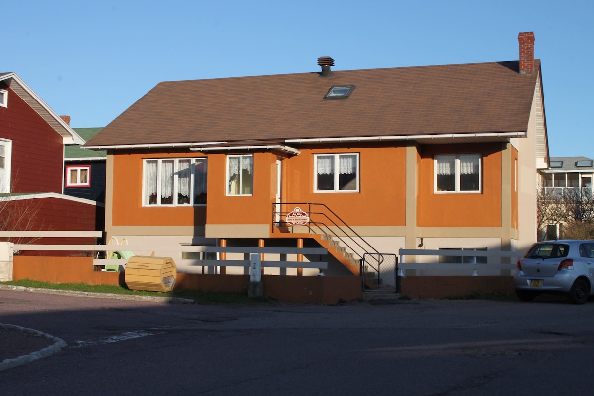 A single-story house features an orange exterior with white trim and multiple windows dressed in lace curtains. A sloped entrance is accessed by a short set of stairs. Surrounding residential structures and a clear blue sky are visible in the background.