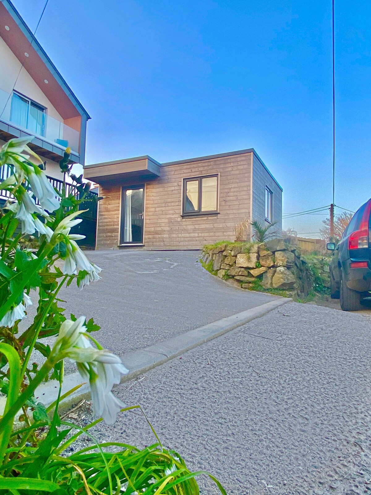 A modern detached unit is positioned at the end of a paved driveway, with a landscaped front area featuring white flowers. The structure boasts large windows and a well-defined entryway. A parked vehicle is visible nearby, with a background of a clear blue sky.