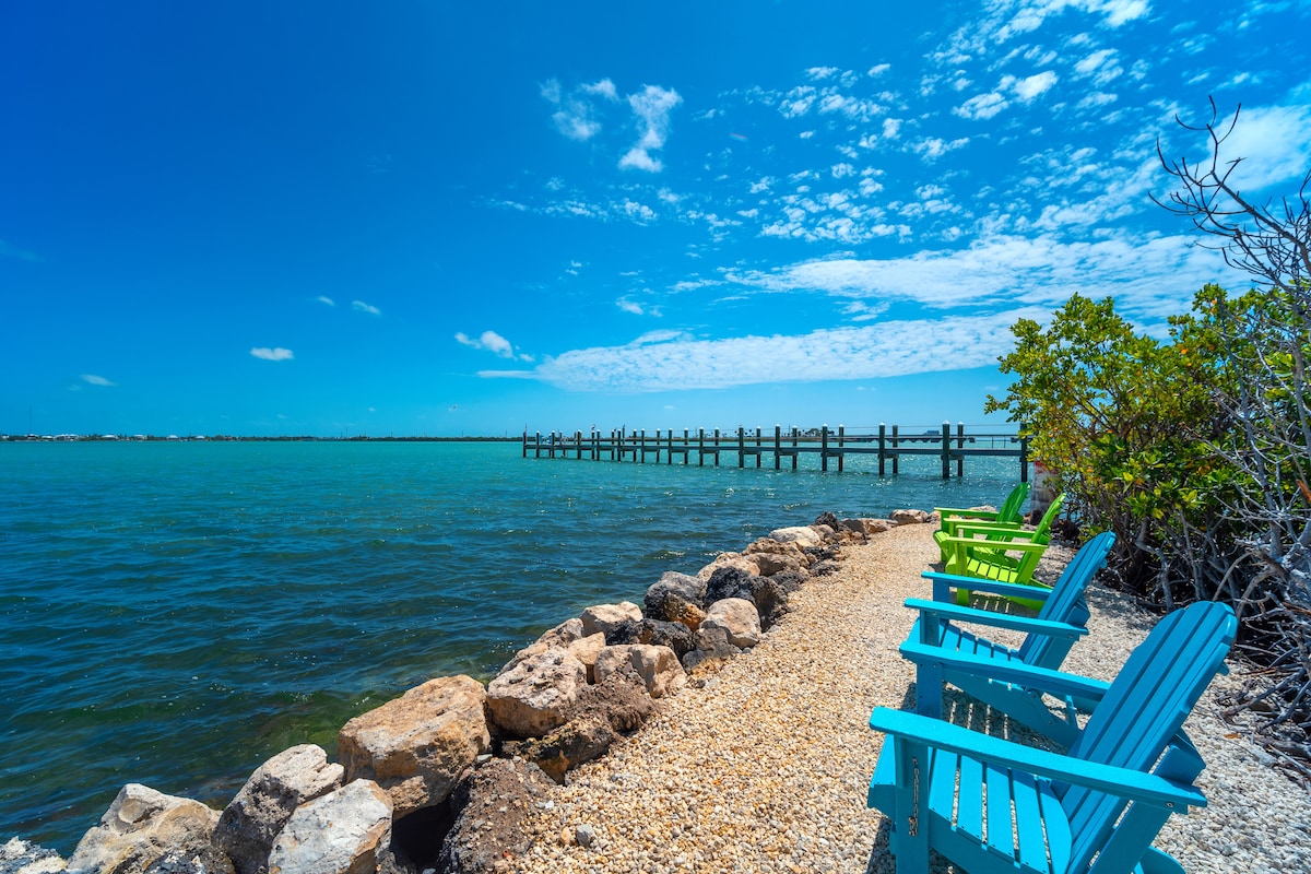 A rocky shoreline is lined with colorful adirondack chairs, offering a perfect view of the clear blue water. A wooden pier extends into the water, accompanied by a backdrop of a bright blue sky with scattered clouds.