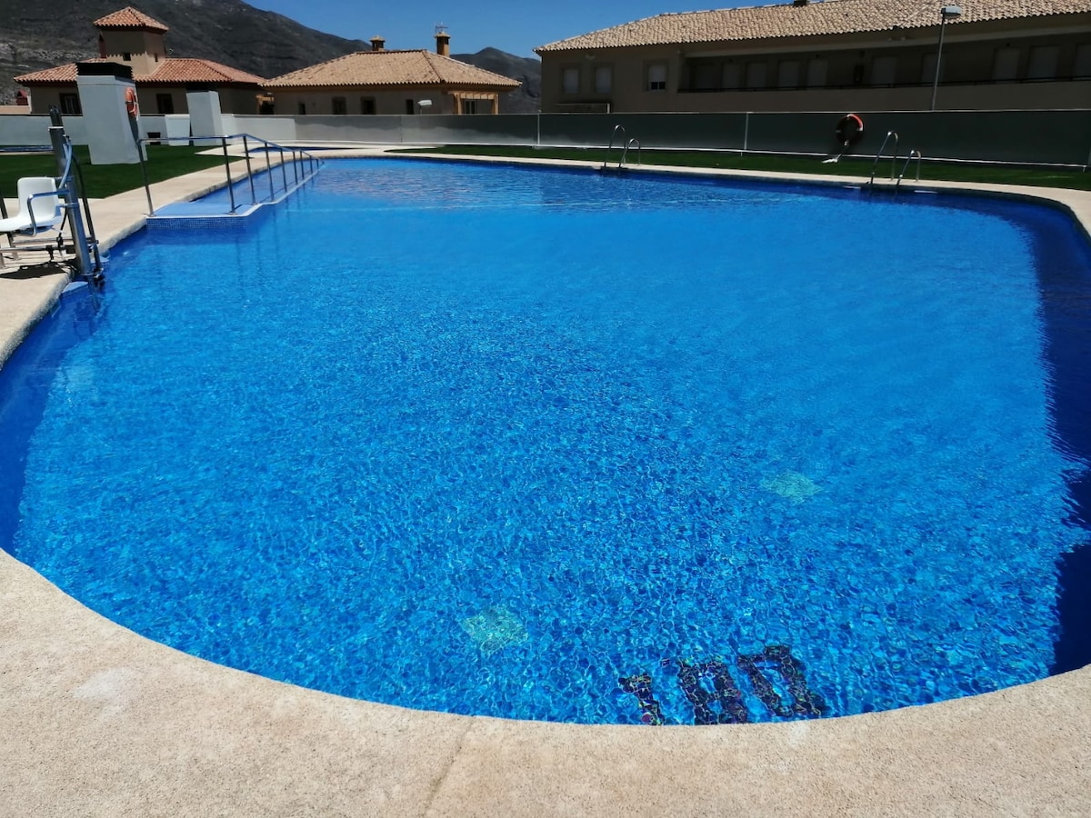 A large outdoor swimming pool is framed by a smooth concrete deck. The water's vibrant blue hue reflects sunlight, creating a serene atmosphere. Nearby, several white lounge chairs are positioned for relaxation. The backdrop features residential buildings with terracotta roofs.