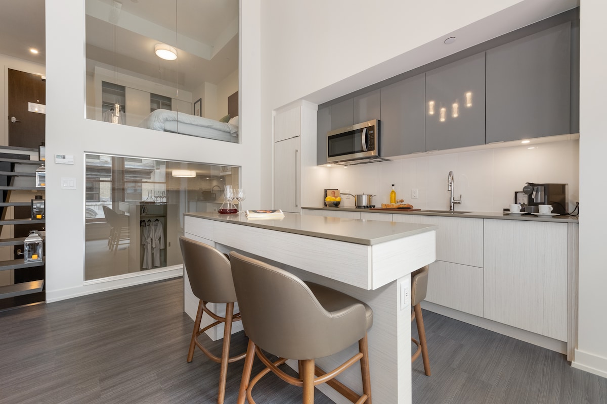 A modern kitchen area is presented with sleek cabinetry and stainless steel appliances. A sizable island with seating for four enhances usability. The dining space provides a view of an elevated sleeping area through glass walls, allowing ample light to fill the open layout.