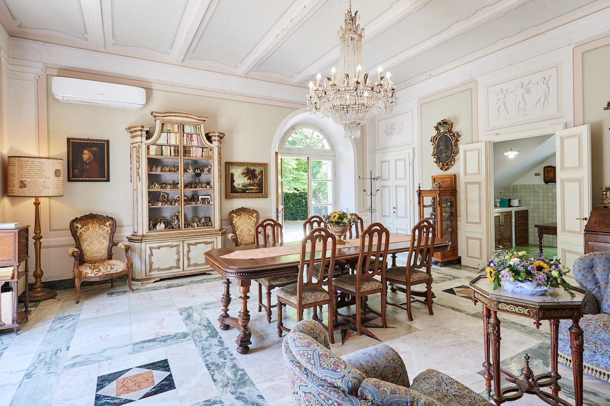 A spacious dining area features a long wooden table surrounded by elegant chairs, with a large chandelier hanging above. Vintage furniture and decor items are showcased in a cabinet, complemented by light streaming through an archway that leads to the kitchen.