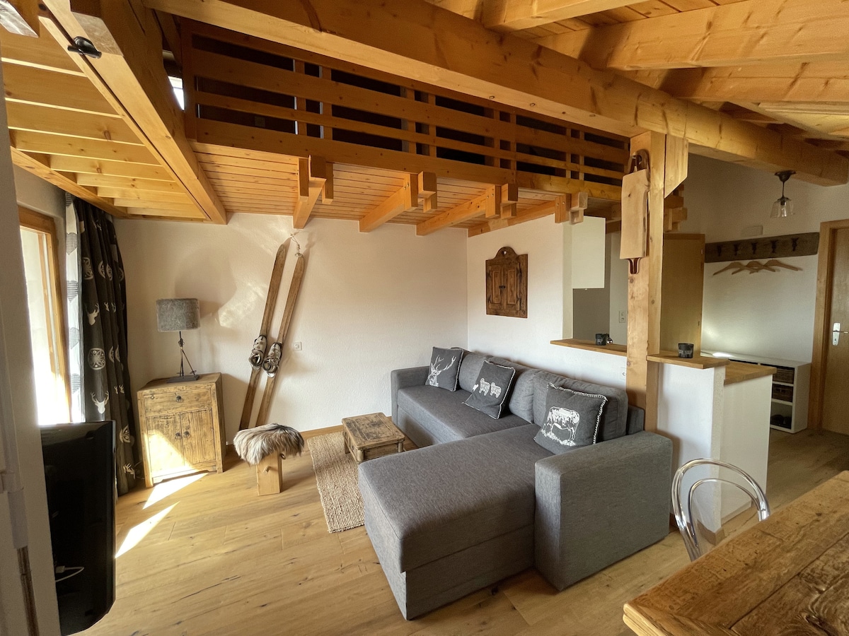 The living area features a spacious sectional sofa arranged around a wooden coffee table. Exposed wooden beams are highlighted in the ceiling. Natural light enters through large windows, while wooden furnishings add a rustic charm to the space.