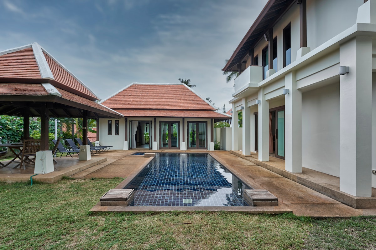 A serene outdoor space is highlighted by a private swimming pool framed by a well-maintained garden. Two separate buildings are visible, showcasing a large covered pool sala and a structured pathway leading towards the lush surroundings. Large glass doors provide a view into the living area.