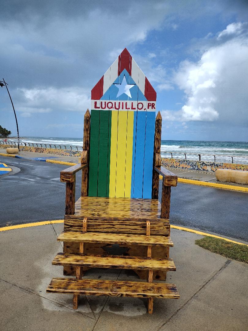 A large, colorful wooden chair is positioned at the beach, adorned with the text 'Luquillo, PR.' The chair features vibrant horizontal stripes of blue, green, and yellow, set against a backdrop of a stormy sky and ocean waves. A concrete walkway leads towards the sea.