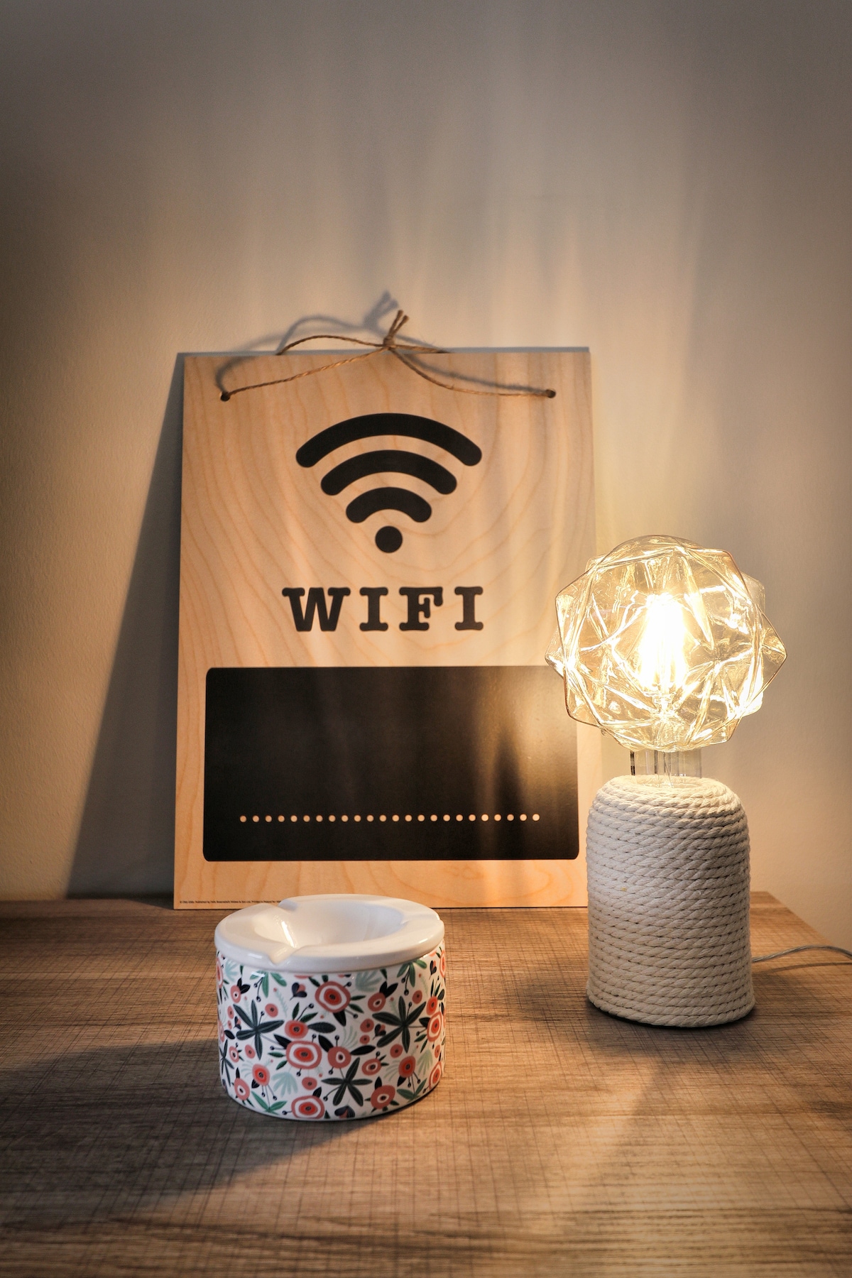 A wooden sign displaying the WiFi symbol is positioned on a textured surface. A decorative candle holder with a floral pattern sits beside a glowing lamp with a geometric design, creating a warm ambiance.