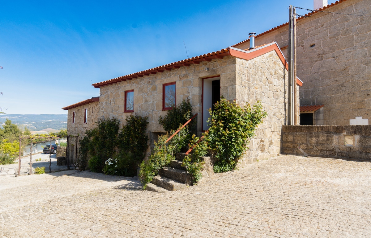 A stone building with a rustic facade is set against a clear blue sky. The entrance features steps lined with greenery, and several windows provide natural light. The surrounding area is made of cobblestones, contributing to the charming village ambiance.