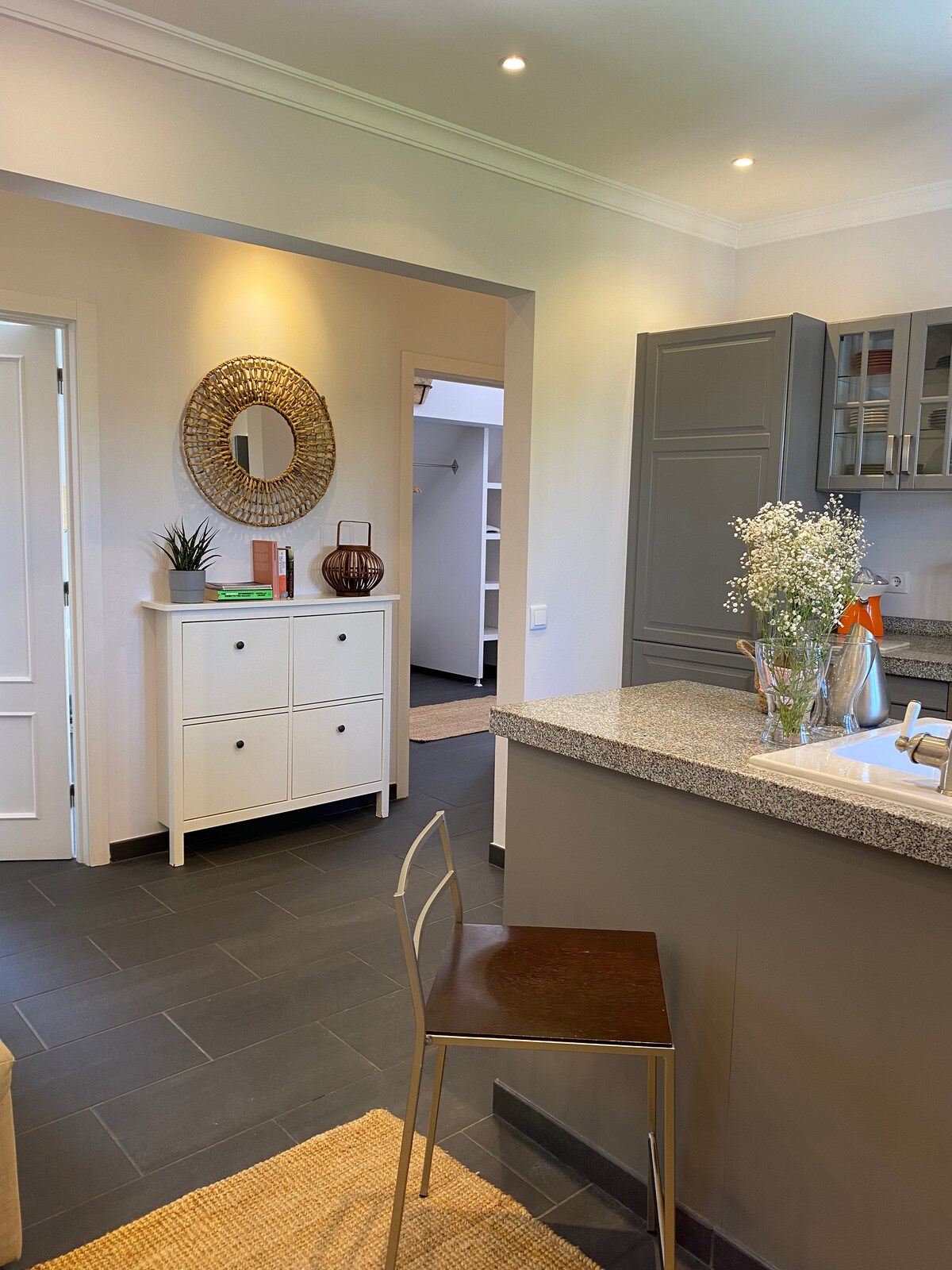 The kitchen area features granite countertops and modern cabinetry. A single metal chair is positioned near the counter. Decor elements, including a decorative mirror and a vase with flowers, add a touch of style. Soft lighting enhances the open and airy feel of the space.