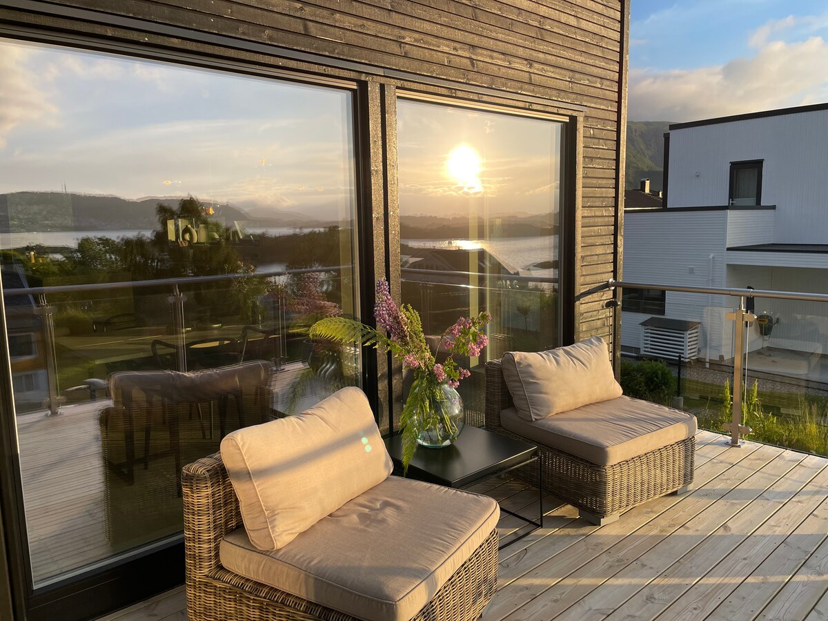 A serene outdoor seating area is showcased, featuring two comfortable lounge chairs with soft cushions. A small table sits between them, adorned with a vase of flowers. Large glass doors reflect the sunset, enhancing the space with a warm glow and offering views of the landscape.