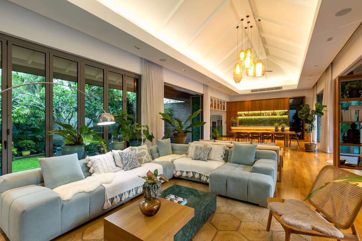A spacious living area features a sectional sofa adorned with cushions, arranged around a coffee table. Natural light floods in through large glass doors that open to a lush garden. Elegant pendant lights hang from a high ceiling, complementing the warm wooden flooring.