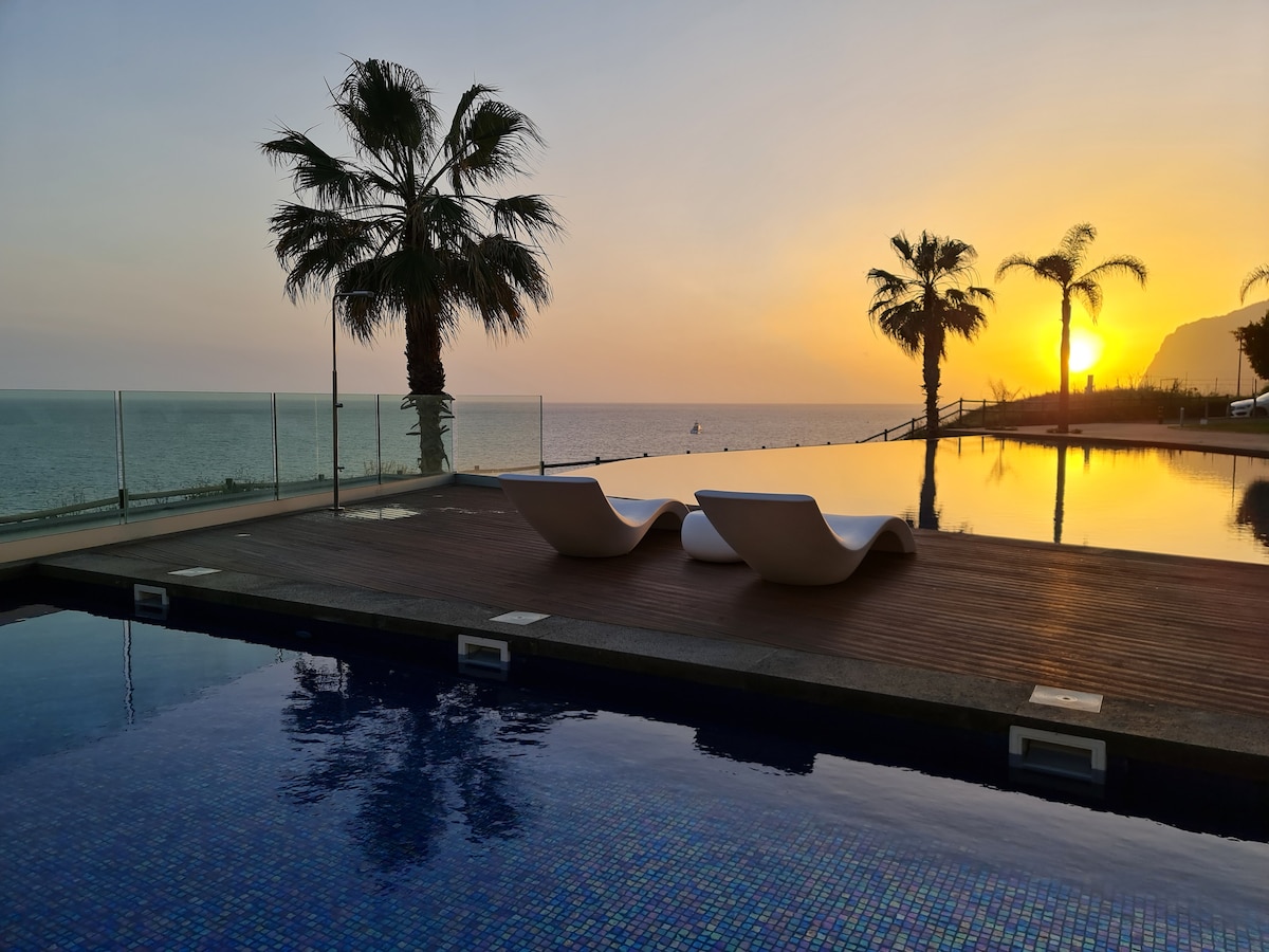 A serene outdoor area features two lounge chairs positioned by an infinity pool, overlooking the ocean. Palm trees add a tropical touch, while a sunset casts warm hues across the sky, creating a peaceful atmosphere.