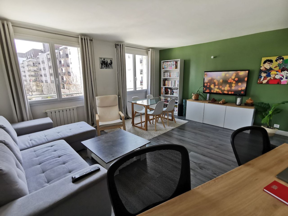 The living area features a spacious grey couch and a light-colored armchair, complemented by a modern coffee table. A dining table with four chairs is positioned near large windows, providing ample natural light. A wall-mounted TV and a bookshelf filled with books add character to the room.