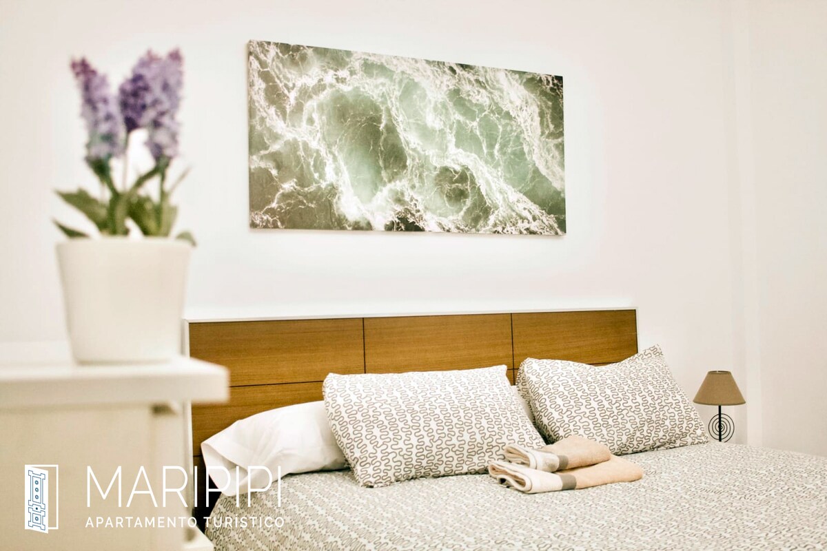 A cozy sleeping area features a wooden headboard with patterned bedding, complemented by soft, neutral colors. Above the bed, an artwork depicting gentle ocean waves adds a calming element. A small lamp provides additional light, while a potted plant adds a touch of greenery.